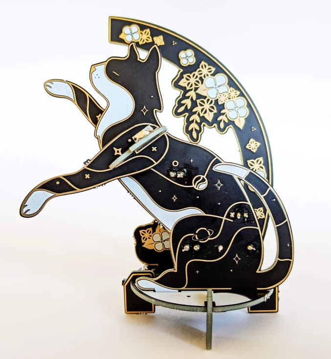 Lötbausatz aus schwarzer Platine in Katzenform mit LEDs, Sternmotiven und floralen PCB-Elementen im Hintergrund.