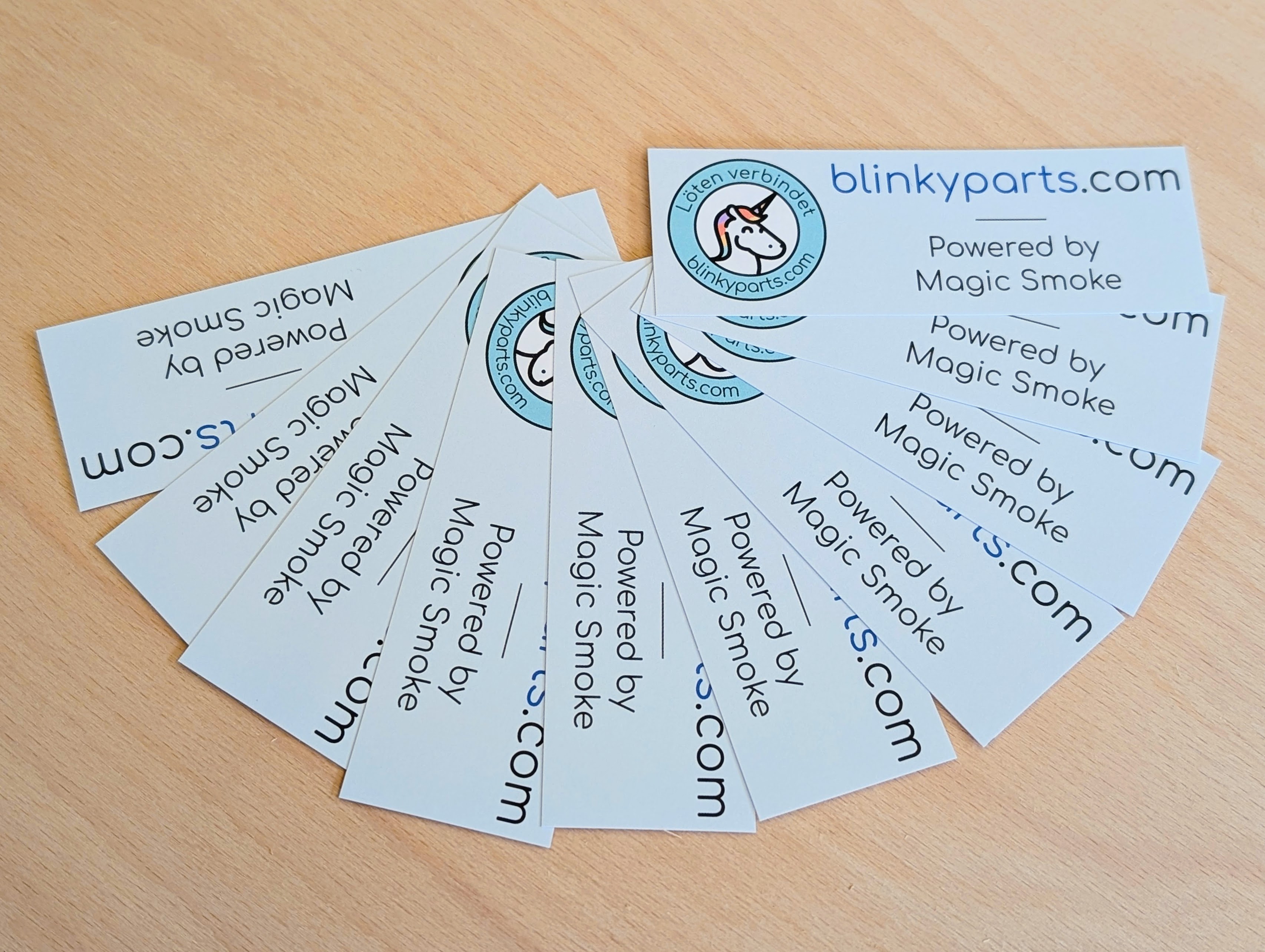 Blinkyparts logo sticker 10-pack – Powered by Magic Smoke 10 Blinkyparts logo stickers fanned out on a table, showing the company logo with web address and slogan ‘Powered by magic smoke’ for electronics enthusiasts