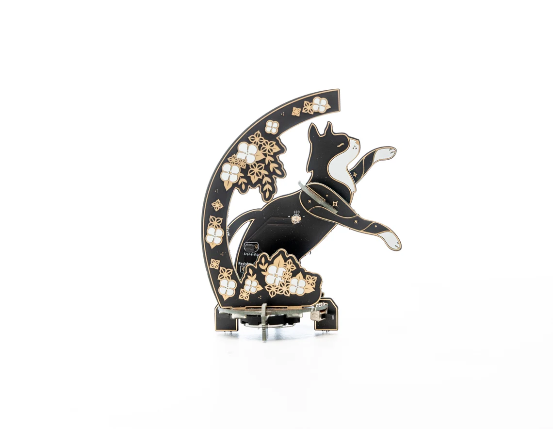 Lötbausatz aus schwarzer Platine in Katzenform mit LEDs, Sternmotiven und floralen PCB-Elementen im Hintergrund.
