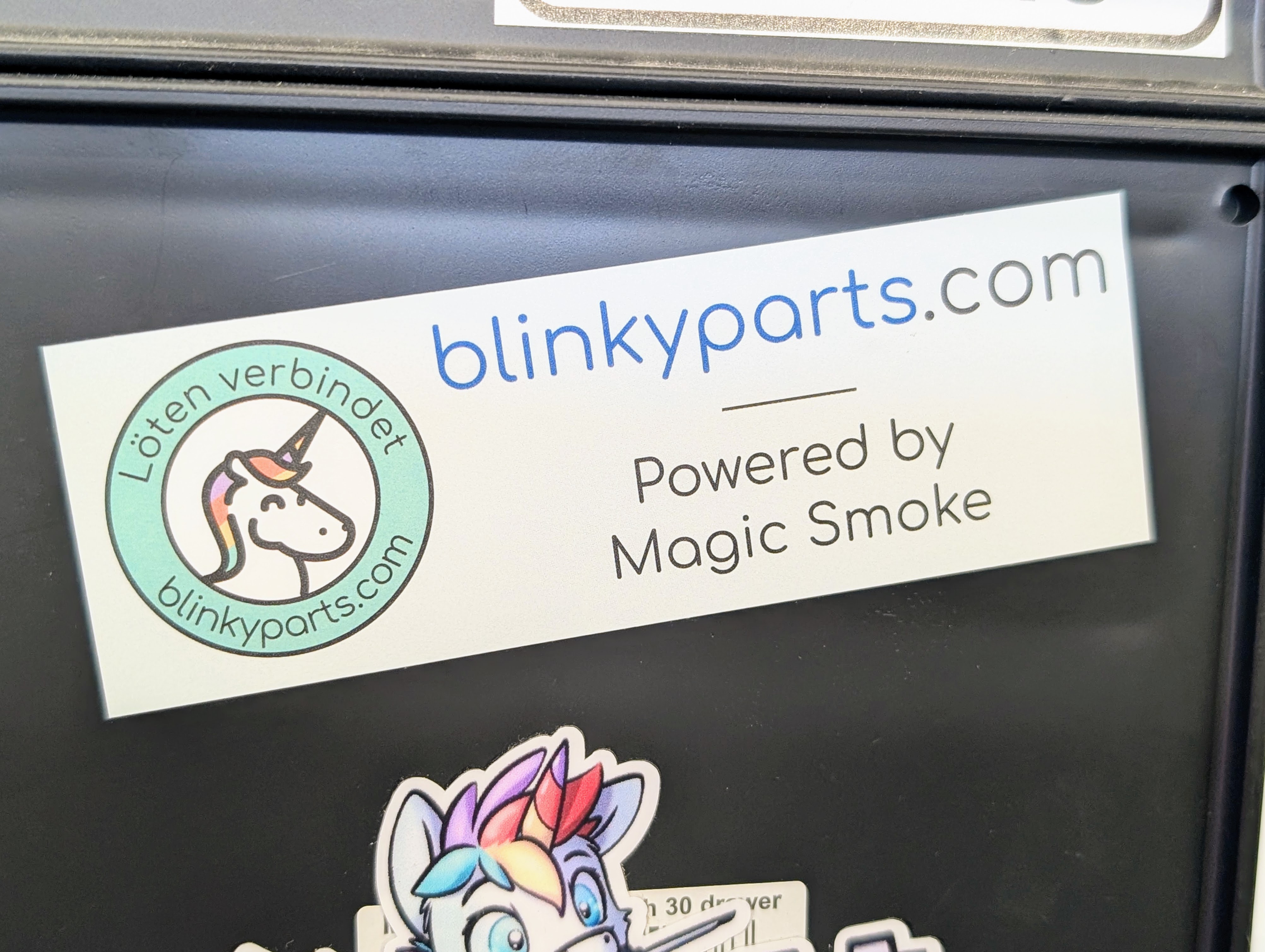 Blinkyparts logo sticker on dark background - contrast view Blinkyparts logo sticker affixed to dark background, showing optimal visibility of the white logo with web address and 'Powered by magic smoke'