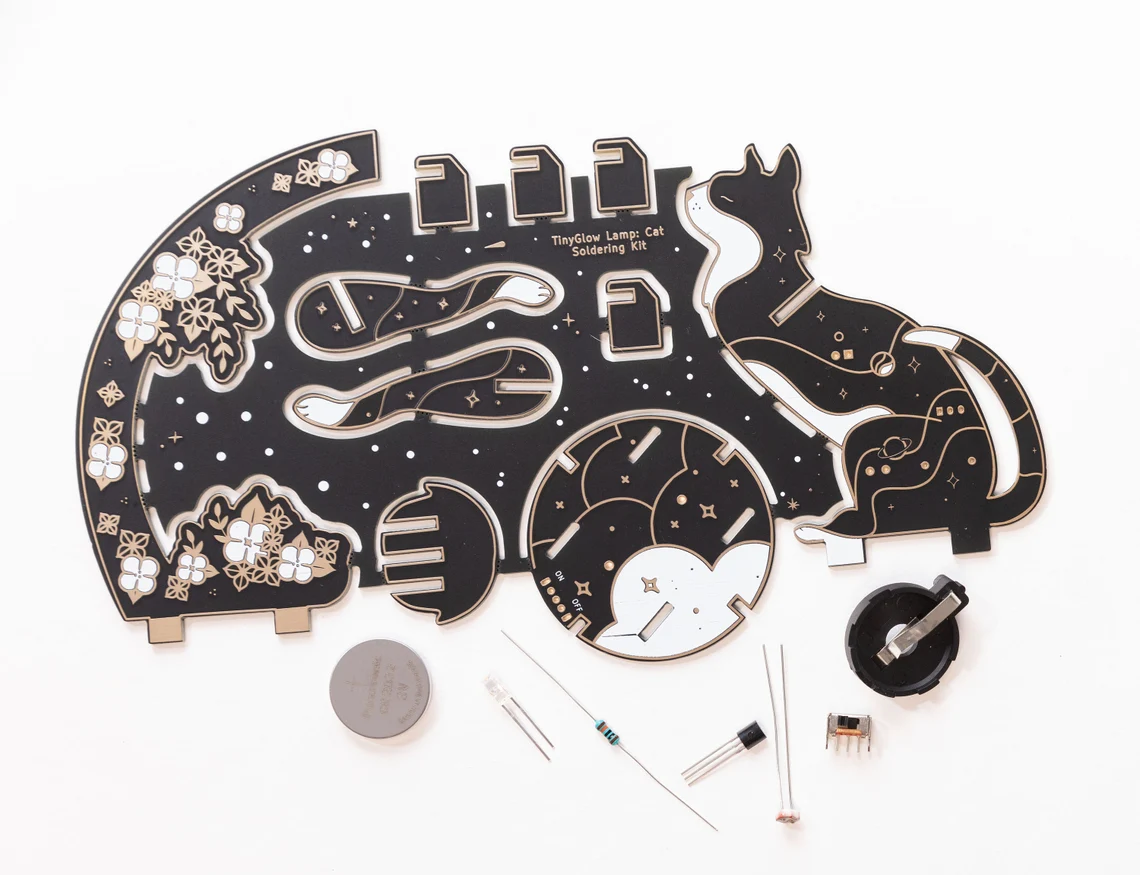  Lötbausatz mit schwarzer PCB-Katzenform, Elektronikbauteilen und dekorativen Elementen zum Aufbau einer Leuchtlampe.