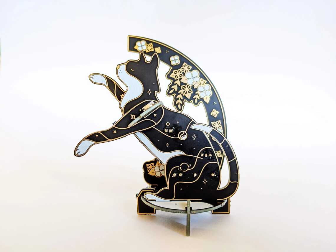 Cat Lamp Soldering Kit Lötbausatz aus schwarzer Platine in Katzenform mit LEDs, Sternmotiven und floralen PCB-Elementen im Hintergrund.